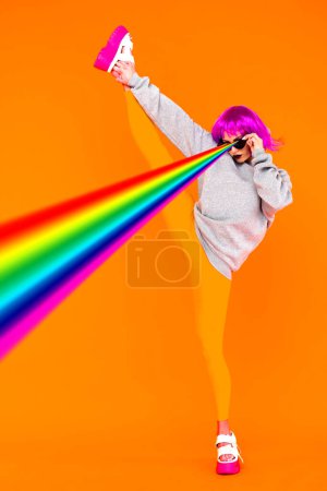 Foto de Una chica lesbiana con una peluca rosa y gafas de sol dispara desde sus ojos láseres de arco iris de pie sobre un fondo naranja, levantando su pierna - Imagen libre de derechos