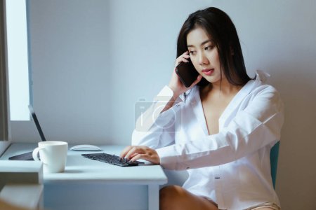 Foto de "La nueva normalidad. Una empresaria está usando su teléfono y su computadora para trabajar para una empresa. a través de Internet en su escritorio en casa." - Imagen libre de derechos
