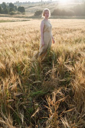 Photo for Young pretty girl in yellow dress stands at field of ears in rays of rising sun. grove and village in background - Royalty Free Image