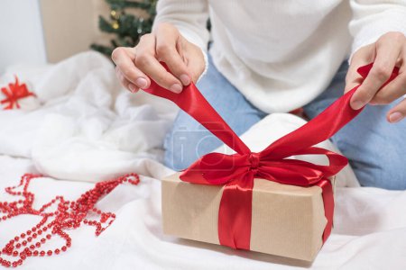 Foto de Felices Fiestas. Las manos femeninas desatan un lazo en una caja de regalo para Navidad en una cama blanca. - Imagen libre de derechos