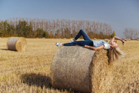 Téléchargez les photos : Belle villageoise posant en jeans sur une balle de foin dans un champ - en image libre de droit