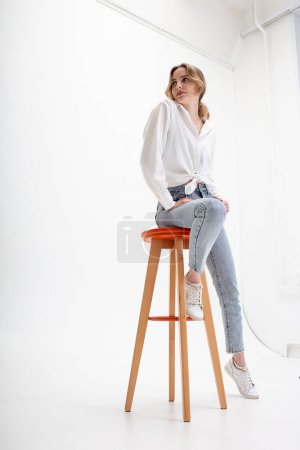 Foto de "joven mujer caucásica pensativa posando en camisa y jeans azules, sentado en el taburete" - Imagen libre de derechos