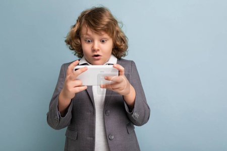 Photo for Photo shot of Handsome amazed emotional child boy with curly hair wearing grey suit holding and using phone isolated over blue background looking at smartphone playing games - Royalty Free Image