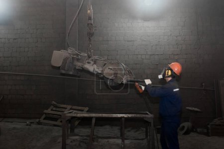 Foto de "Un trabajador con un mono, un casco protector y un respirador controla el equipo pesado para la limpieza de tubos de hormigón de hierro fundido en el taller de una planta industrial" - Imagen libre de derechos