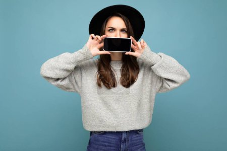Foto de Foto de Hermosa joven positiva con sombrero negro y suéter gris sosteniendo el móvil mostrando el teléfono inteligente aislado en el fondo mirando a la cámara - Imagen libre de derechos