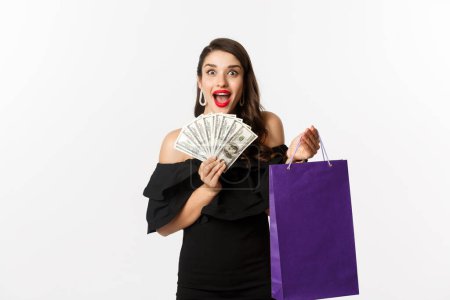 Téléchargez les photos : Image de belle femme excitée en robe noire faisant du shopping, tenant sac et dollars, debout sur fond blanc - en image libre de droit