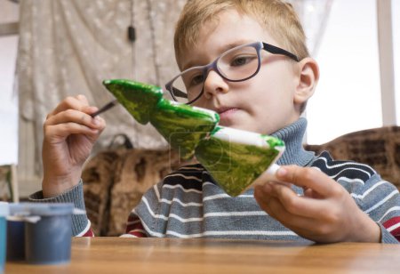 Téléchargez les photos : "Un garçon de sept ans peint un sapin de Noël en polystyrène vert en préparation de Noël. Idées cadeaux de Noël faits à la main." - en image libre de droit