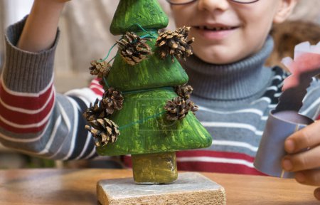 Téléchargez les photos : "Un petit garçon décore un sapin de Noël fait à la main avec des cônes de pin et en même temps joue drôle." - en image libre de droit