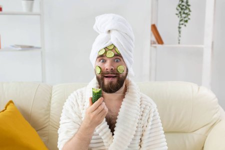 Téléchargez les photos : "Homme barbu drôle tenant des tranches de concombre sur le visage. Spa à la maison, soins du corps et de la peau pour le concept masculin." - en image libre de droit