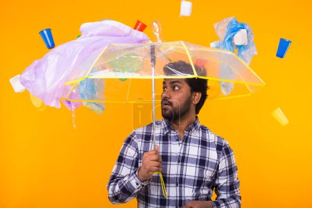Photo for "Problem of trash, plastic recycling, pollution and environmental concept - Surprised man is standing under trash with umbrella on yellow background" - Royalty Free Image