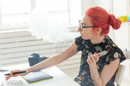 Téléchargez les photos : Créatif, graphiste, concept de personnes - jeune femme créative fumant une vape tout en travaillant avec une tablette graphique - en image libre de droit