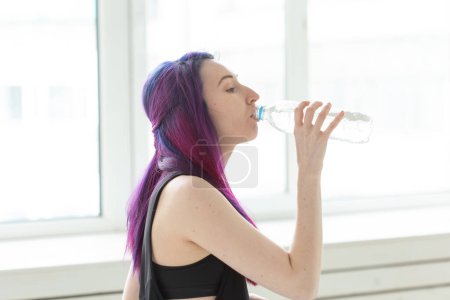 Téléchargez les photos : "Jeune fille hipster avec des cheveux colorés boit de l'eau d'une bouteille après un entraînement physique intense dans la salle de gym. Le concept de soif et équilibre hydrique du corps
." - en image libre de droit