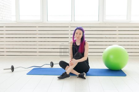 Photo for "Young mixed race girl hipster with purple hair is sitting on the sport mat next to the barbell and with the fitball in the gym. Concept of regular exercise and fitness." - Royalty Free Image