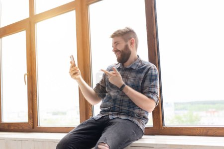 Téléchargez les photos : Mignon jeune gars positif hipster avec une barbe est assis sur le rebord de la fenêtre par la grande fenêtre et parler sur la communication vidéo à l'aide d'un smartphone et Internet haut débit sans fil
. - en image libre de droit