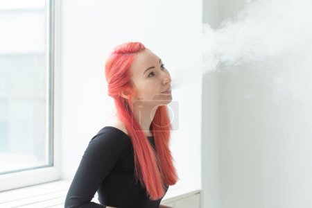Foto de "Vape, adicción y concepto de personas - mujer con el pelo rojo está fumando vapor
" - Imagen libre de derechos