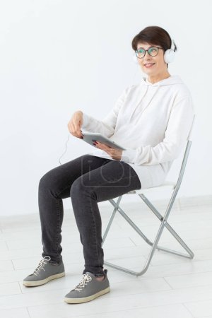 Téléchargez les photos : Charmante femme mince d'âge moyen dans des lunettes et un pull est assis sur une chaise et écoute sa radio en ligne préférée avec l'aide d'un casque et Internet sans fil sur un fond blanc - en image libre de droit