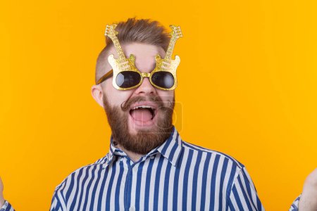 Photo for Positive young man with glasses in the form of guitars rejoices against a yellow background. The concept of celebration and parties. - Royalty Free Image