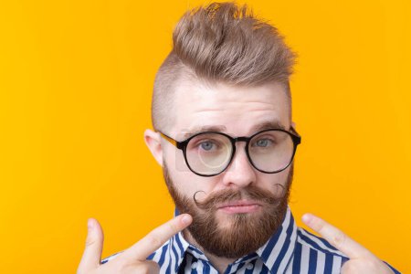 Photo for Charming confident young fashion hipster man with glasses and a beard shows on himself posing over a yellow background. Place for advertising. The concept of self-confidence and success. - Royalty Free Image