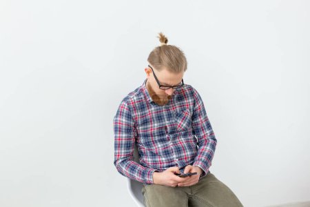 Téléchargez les photos : Jeune homme hipster barbu tenant son téléphone portable sur le fond blanc - en image libre de droit