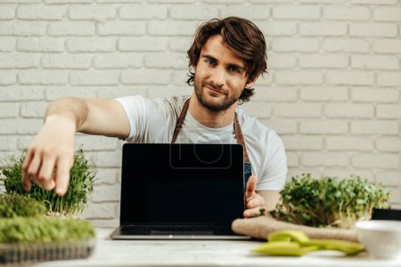 Photo for Man farmer shows black screen of laptop and sits at the table with sprouts - Royalty Free Image