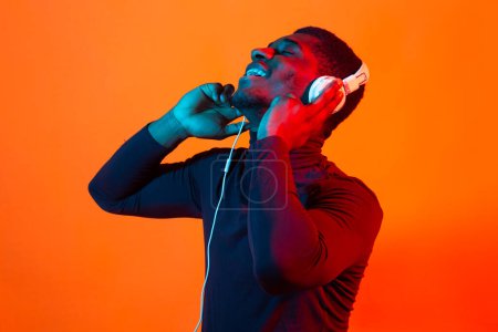 Photo for "African american young man listening to music online dancing and singing with headphones, neon light. Music and technology concept." - Royalty Free Image