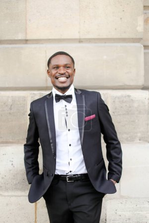 Foto de Afro americano sonriente hombre vistiendo traje oscuro y de pie cerca de la pared al aire libre. - Imagen libre de derechos