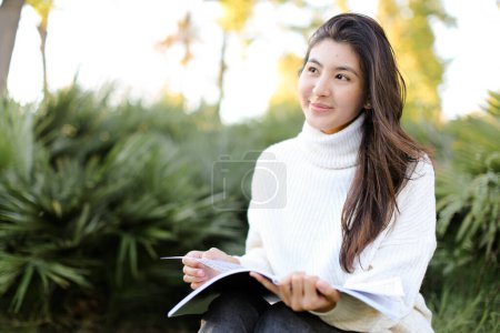Photo for Chinese smiling girl sitting on stump in park and reading papers. - Royalty Free Image