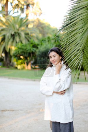 Foto de Chica linda coreana en suéter blanco de pie cerca de hoja de palma. - Imagen libre de derechos