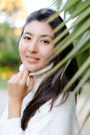 Foto de Retrato de niña bonita china con suéter blanco de pie cerca de la hoja de palma. - Imagen libre de derechos