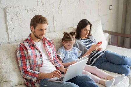 Photo for Father, mother and daughter using electronic devices sitting on sofa at living room. - Royalty Free Image