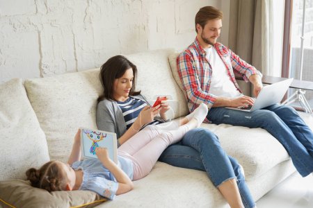 Téléchargez les photos : Père, mère et fille utilisant des appareils électroniques assis sur le canapé dans le salon. - en image libre de droit