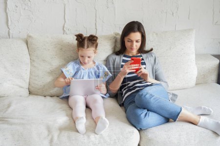 Photo for Mother and daughter using electronic devices sitting on sofa at living room. - Royalty Free Image