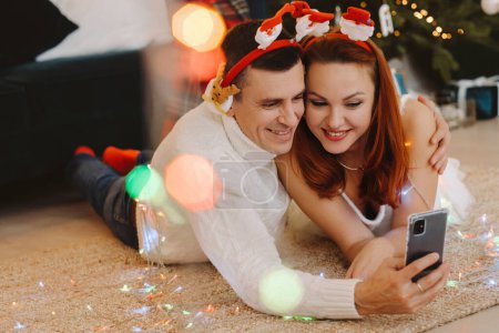 Foto de Una pareja casada feliz está tirada en el suelo en casa cerca del árbol de Navidad y tomando fotos de sí mismos - Imagen libre de derechos
