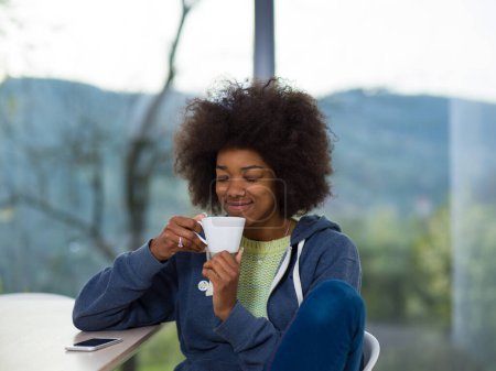 Foto de Mujer negra bebiendo café y usando un teléfono móvil en casa - Imagen libre de derechos
