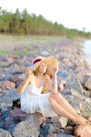 Photo for Young pretty woman in hat sitting on shingle beach - Royalty Free Image