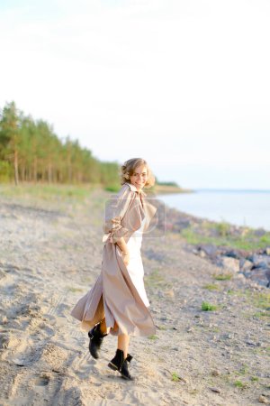 Téléchargez les photos : Vue arrière de la jeune femme blonde marchant sur la plage de bardeaux et portant un manteau d'été. - en image libre de droit
