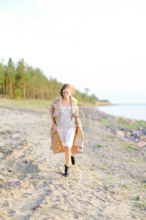 Téléchargez les photos : Jeune belle femme marchant sur la plage de bardeaux et portant un manteau d'été. - en image libre de droit