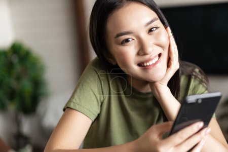 Téléchargez les photos : Souriant coréen fille regardant heureux, tenant smartphone et assis dans le salon à la maison. Concept de style de vie - en image libre de droit