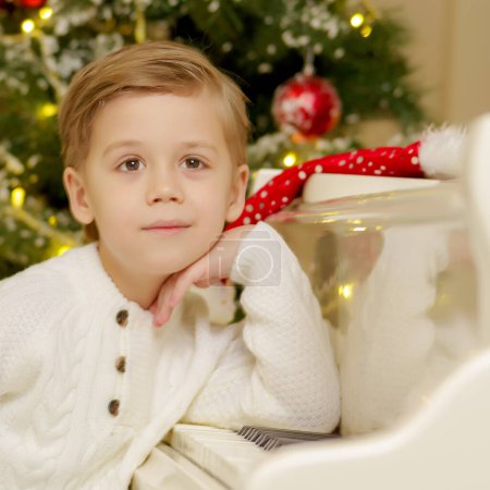Téléchargez les photos : Un petit garçon est assis au piano près du sapin de Noël. - en image libre de droit