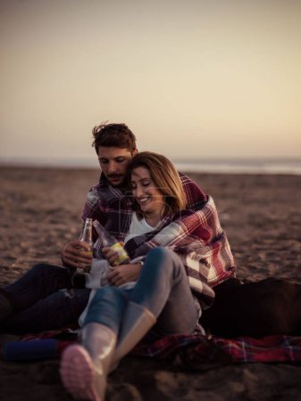 Foto de Amar a la joven pareja sentada en la playa junto a la fogata bebiendo cerveza - Imagen libre de derechos
