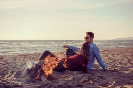 Téléchargez les photos : Jeune couple assis sur la plage à côté de feu de camp boire de la bière - en image libre de droit