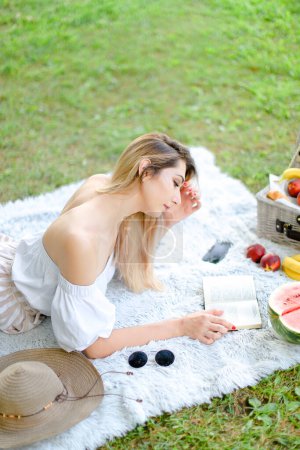 Foto de Joven chica caucásica leyendo libro y acostado sobre cuadros cerca de frutas y sombrero, hierba en el fondo. - Imagen libre de derechos