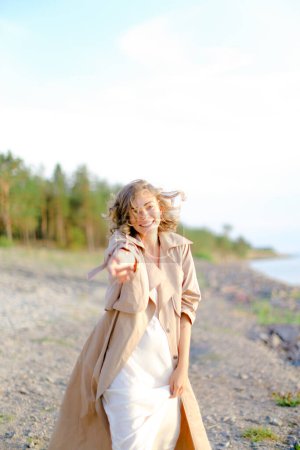 Téléchargez les photos : Jeune jolie femme avec la main tendue sur la plage de bardeaux portant un manteau d'été. - en image libre de droit