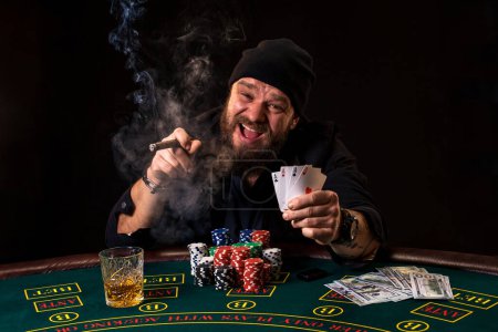 Téléchargez les photos : L'homme joue au poker avec un cigare et un whisky, un homme montre deux cartes à la main, remportant toutes les jetons sur la table avec une épaisse fumée de cigarette
. - en image libre de droit