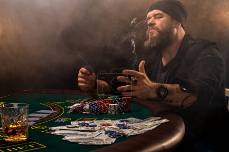 Foto de Hombre barbudo con cigarro y vidrio sentado en la mesa de póquer en un casino. Apuestas, cartas y ruleta
. - Imagen libre de derechos