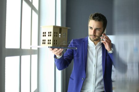 Foto de "Empresario sosteniendo la casa en miniatura en la mano de pie en la oficina
." - Imagen libre de derechos