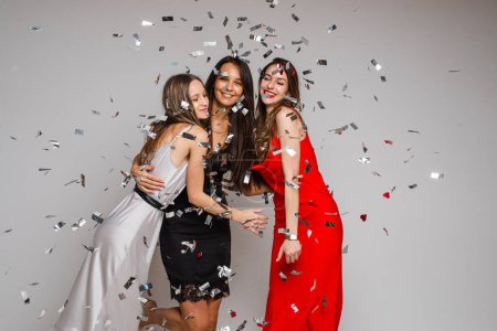 Foto de "Celebración de las jóvenes amigas con vestidos de noche abrazándose bajo confeti de plata en la fiesta de vacaciones en el fondo gris" - Imagen libre de derechos