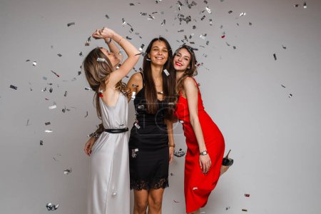 Photo for "Celebrating young girls friends wearing evening dresses hugging under silver confetti on holiday party on gray backdrop" - Royalty Free Image
