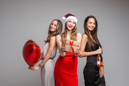 Foto de "Guapas jóvenes amigas en vestidos festivos sosteniendo regalo, globo rojo, champán celebrando la fiesta en el fondo gris" - Imagen libre de derechos