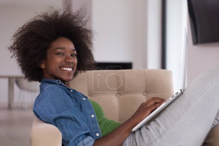 Téléchargez les photos : Femme afro-américaine à la maison dans la chaise en utilisant un ordinateur portable - en image libre de droit